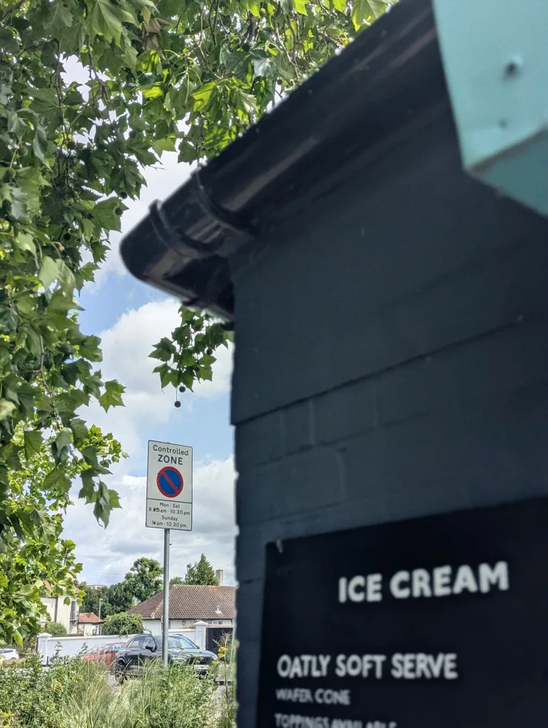 An Oatly soft serve ice cream sign on a dark teal brick building, with a controlled parking zone sign and leafy trees in the background.