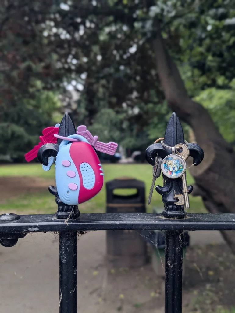 A pink and blue toy and a set of keys with a dog charm hanging on the fleur-de-lis spikes of a black metal fence in a park.