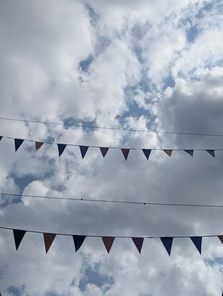 Two strings of multicolored triangular bunting flags against a bright, cloudy blue sky.