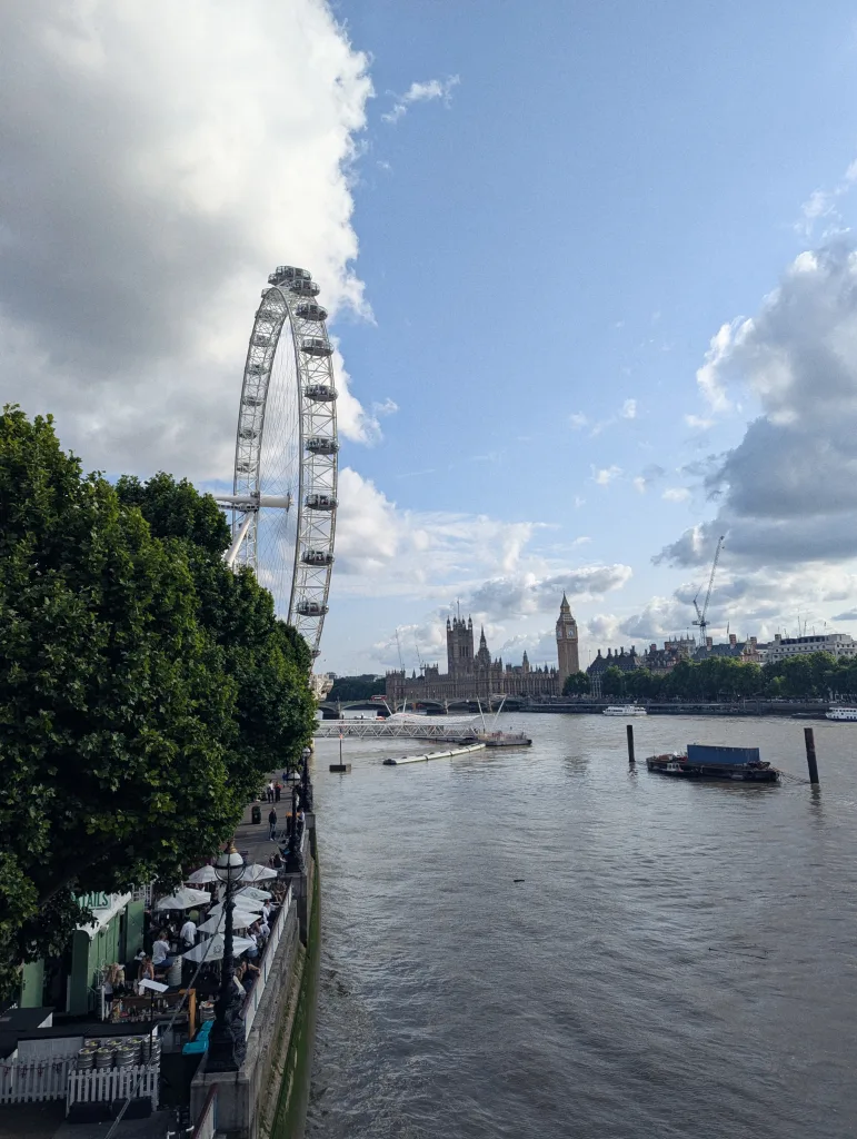 The London Eye towers over the River Thames with Big Ben and the Houses of Parliament in the distance under a bright, cloudy sky.