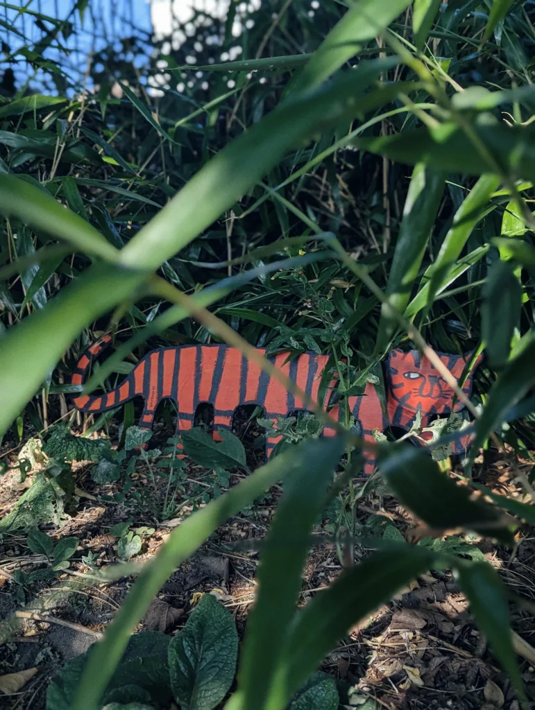 An orange and black striped tiger cutout peeking out from behind dense green bamboo leaves in a garden.