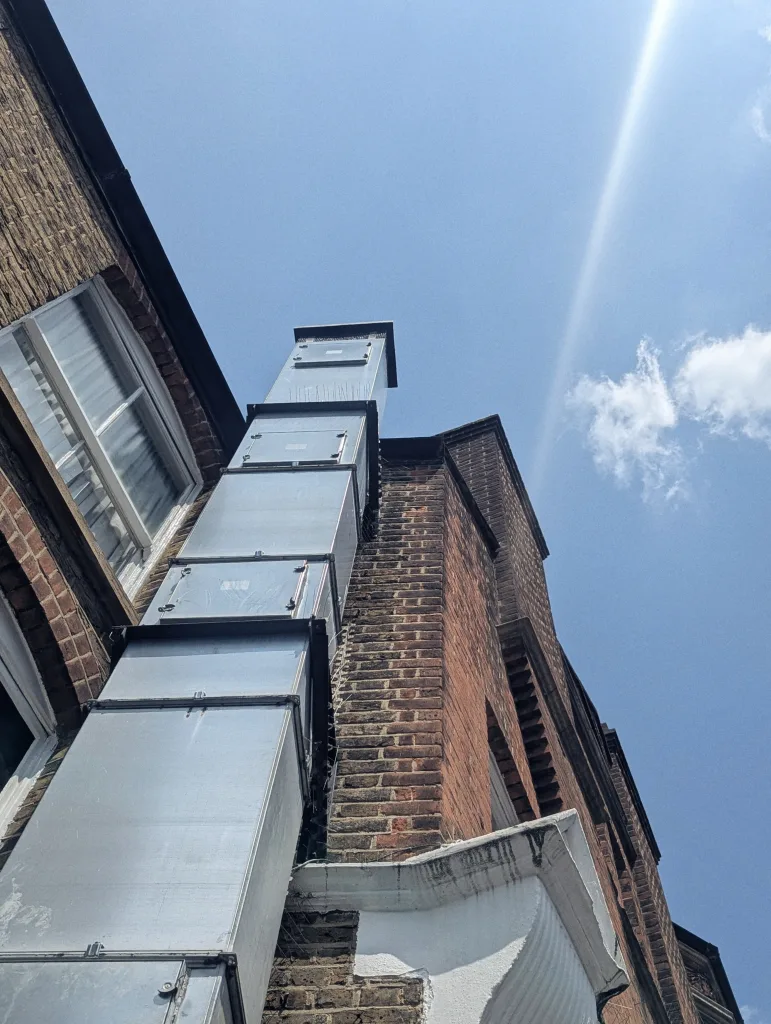 Large rectangular metal ventilation duct running up the side of a brick building against a bright blue sky with a lens flare.