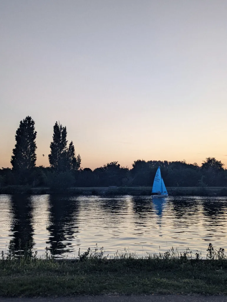 A sailboat with a bright blue sail on a calm river at sunset. Silhouetted tall trees are reflected in the water against a soft orange sky.