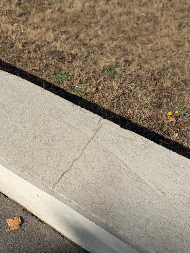 A thin crack across a concrete curb separating a dry brown lawn from asphalt with a single dried leaf.