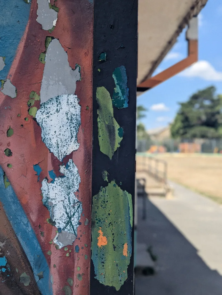 Peeling layers of red, blue, and green paint on a vertical post, with a soft-focus background of a park under a bright blue sky.