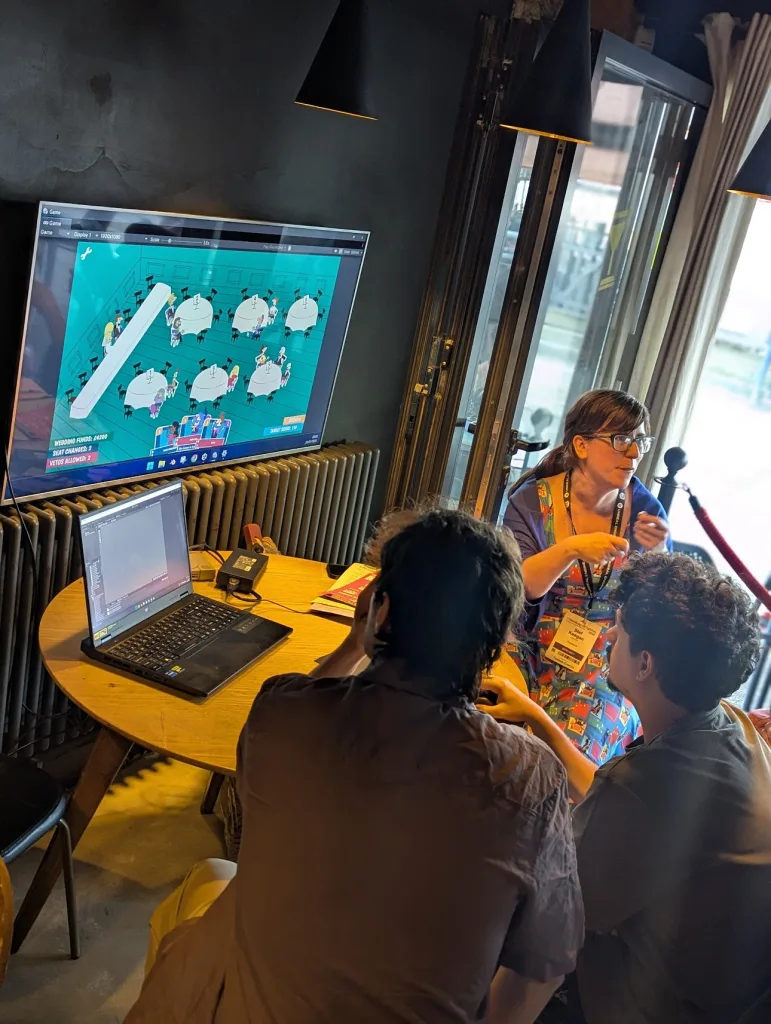 A woman demos a top-down wedding seating game on a large screen to two people at a table with a laptop in a dimly lit room.