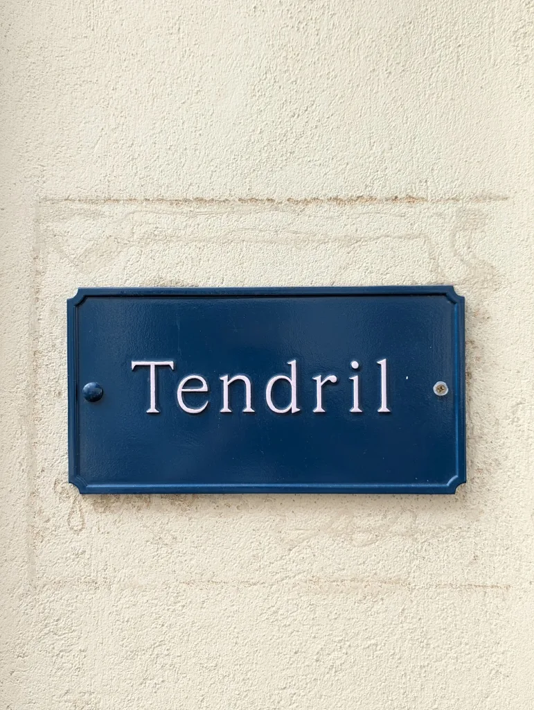 A dark blue rectangular sign with the word Tendril in white serif font, mounted on a textured cream wall.