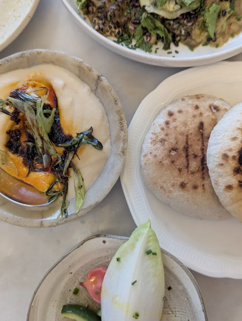 Small plates of creamy dip with chili oil and charred onions, warm pita bread, and fresh endive with pickled vegetables.