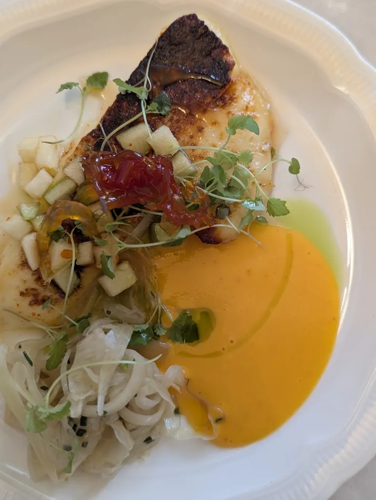 Seared white fish topped with microgreens and diced apple, served with bright orange puree and a fennel salad on a white scalloped plate.