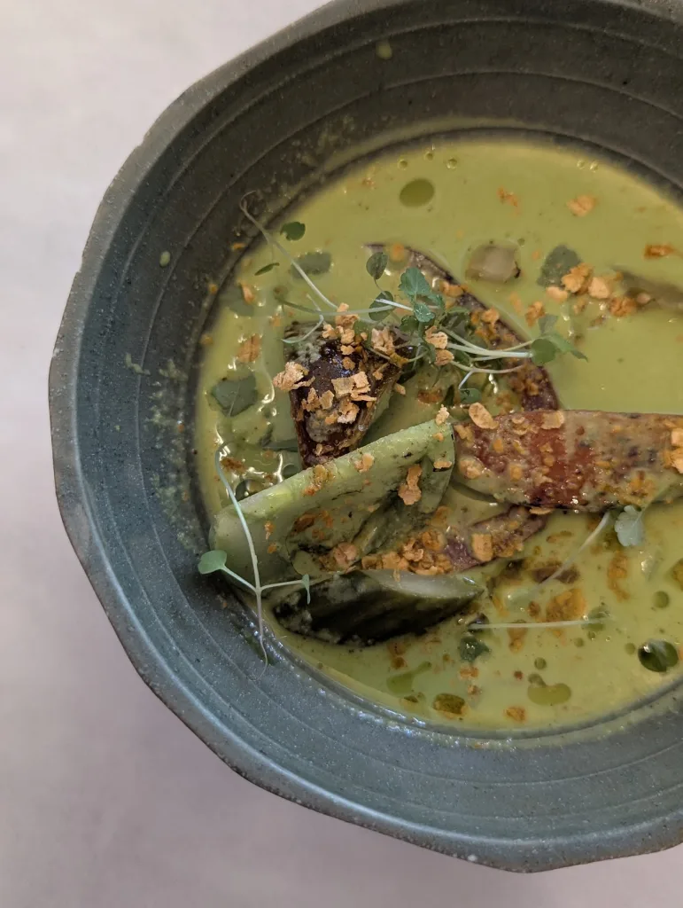 Vibrant green soup in a textured grey ceramic bowl, garnished with charred vegetables, delicate microgreens, and crispy golden toppings.
