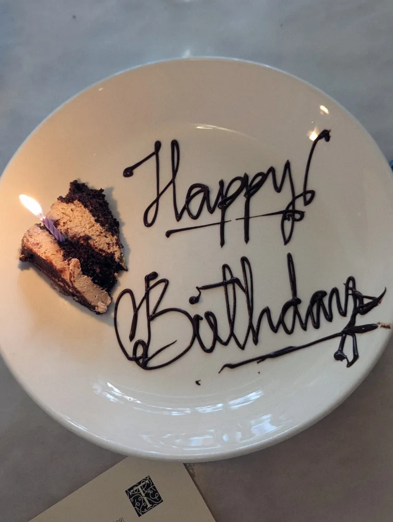 A small slice of chocolate layer cake with a lit purple candle on a white plate with "Happy Birthday" written in cursive chocolate sauce.