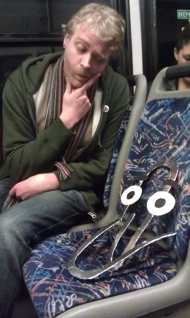 A man sitting on a bus thoughtfully contemplating a large, handmade foil Clippy mascot in the seat next to him.