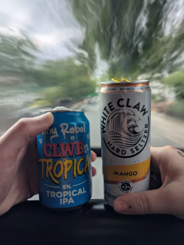 Hands holding cans of Tiny Rebel Tropical IPA and Mango White Claw against a long-exposure motion-blurred background of trees from a moving vehicle.