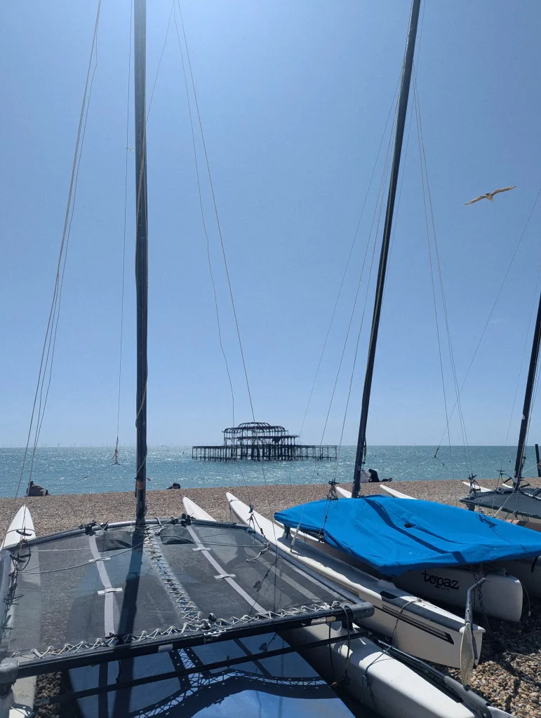 Sailing catamarans on a shingle beach with the skeletal ruins of Brighton's West Pier in the sea under a clear blue sky.