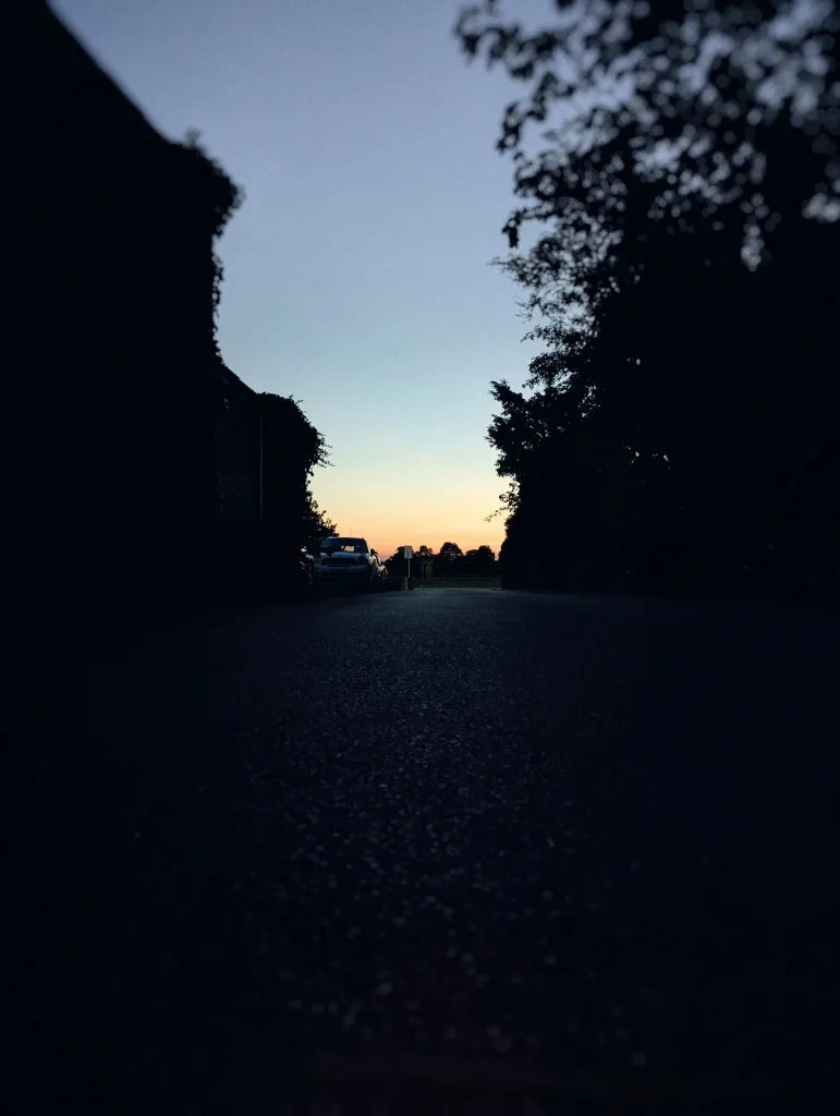 Low-angle view of a tarmac road leading toward a glowing orange and blue sunset horizon, framed by dark silhouetted trees and buildings.