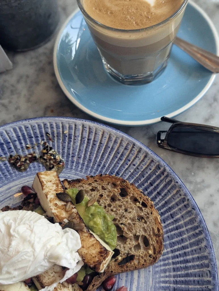 Poached egg, halloumi, and avocado on sourdough toast on a blue plate next to a coffee and sunglasses on a marble tabletop.