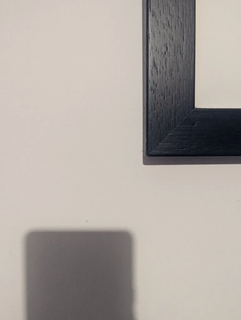 Corner of a black wooden frame on a pale wall with a dark shadow cast in the bottom left corner.