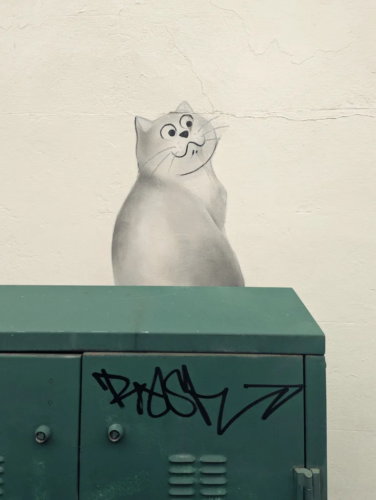 A grey cartoon cat mural peeks over a green metal utility box with black graffiti against a white wall.