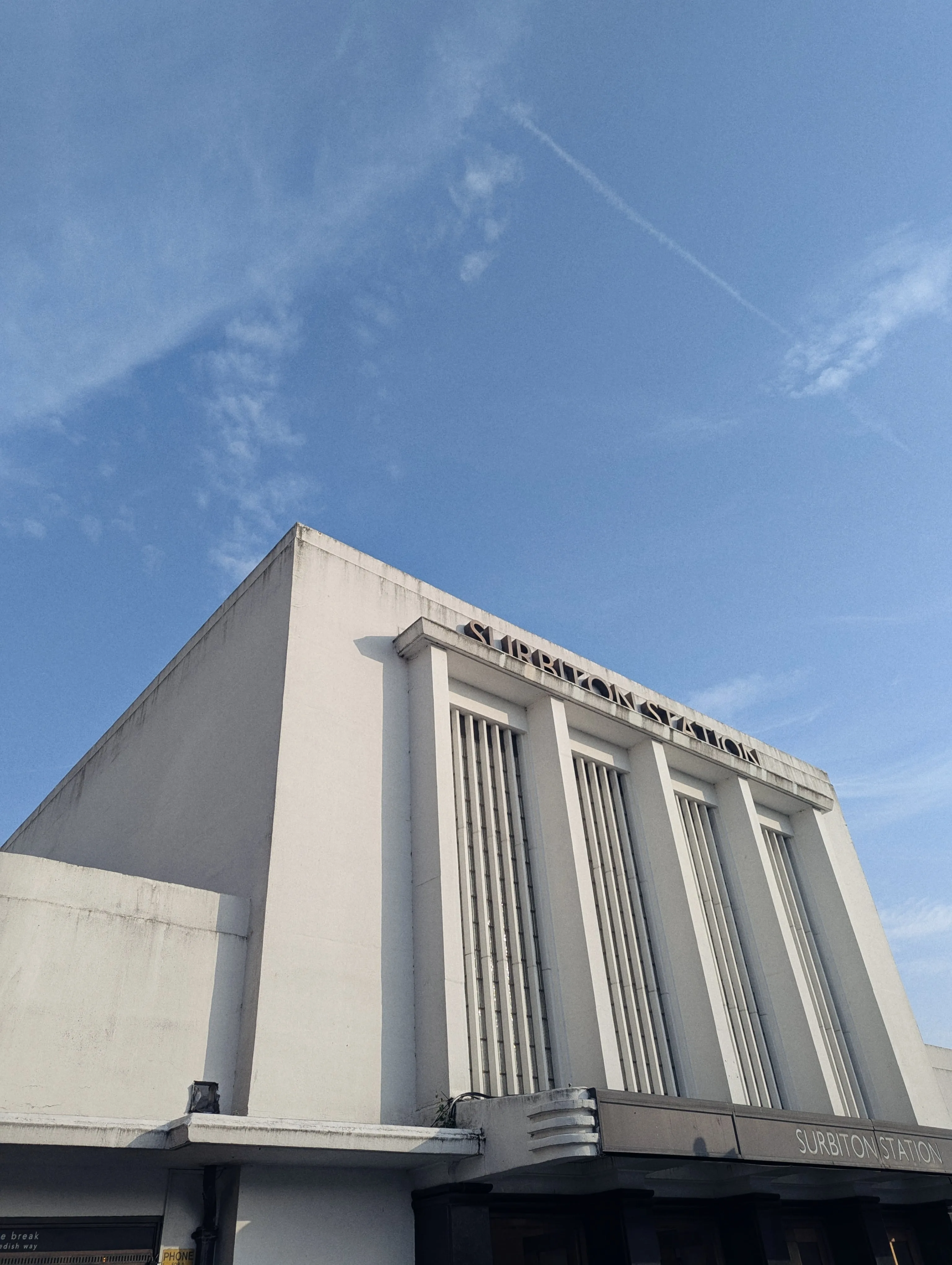 Low angle of the white Art Deco facade of Surbiton Station against a bright blue sky with wispy clouds and a contrail.
