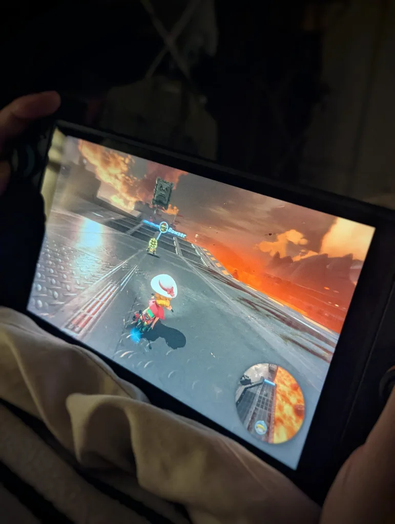 A handheld console playing Mario Kart World on a fiery track, held by a person in a dimly lit room over a soft blanket.