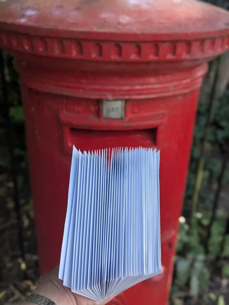 A hand holding a thick fan of light blue envelopes in front of a red British pillar post box.