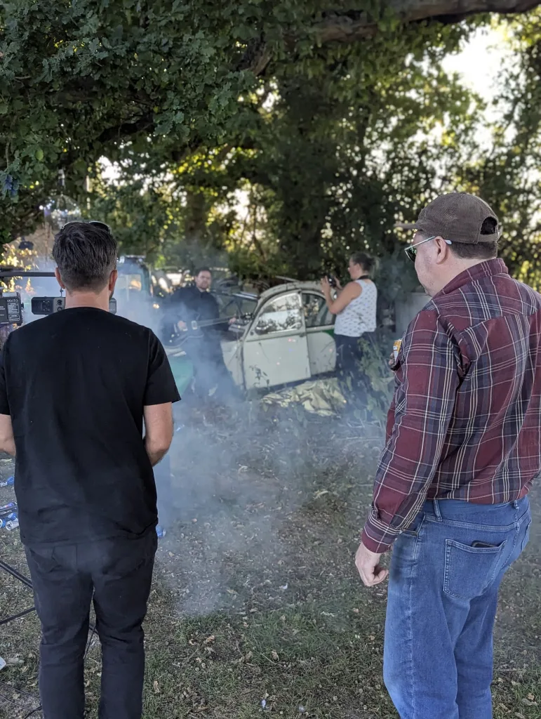 A man plays electric guitar beside a white vintage car in a smoky forest clearing, with a camera operator and onlookers in the foreground.