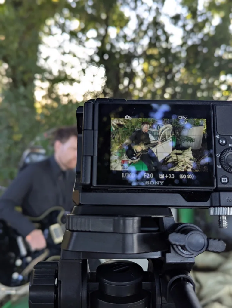 A man playing a guitar viewed through a Sony camera screen on a tripod, with the actual musician blurred in the background of the outdoor setting.