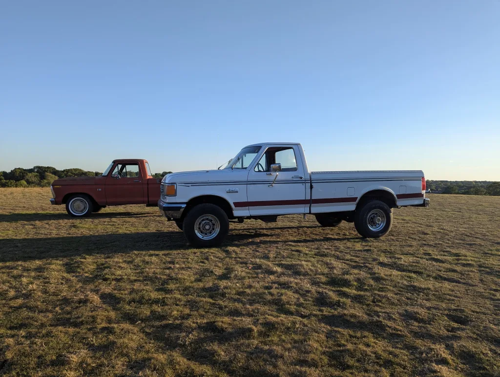 A white Ford F-250 with a red stripe and a vintage red truck parked in a golden grassy field under a clear blue sky at sunset.