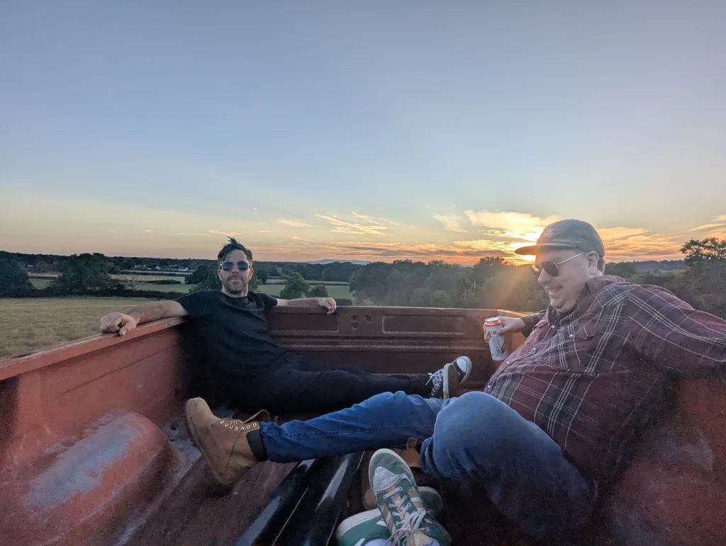 Two men relax in the back of a rusty red pickup truck bed, watching a sunset over a rolling rural landscape.