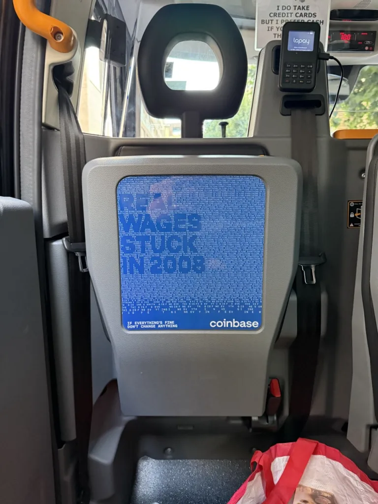 A Coinbase ad on a taxi seat back reads "REAL WAGES STUCK IN 2008" over a blue background of repeating "EVERYTHING IS FINE" text.