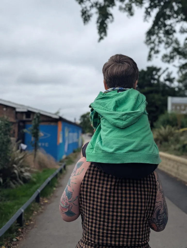 A child in a green hoodie on the shoulders of a person in a checkered shirt with tattooed arms, walking along a path past a blue wall.