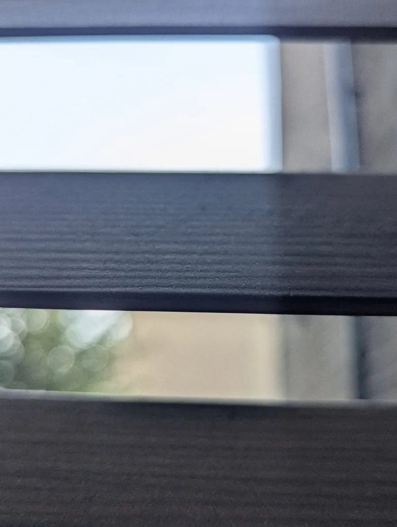 Close-up of dark grey horizontal slats showing a blurry view of bright sunlight and green bokeh through the narrow gaps.