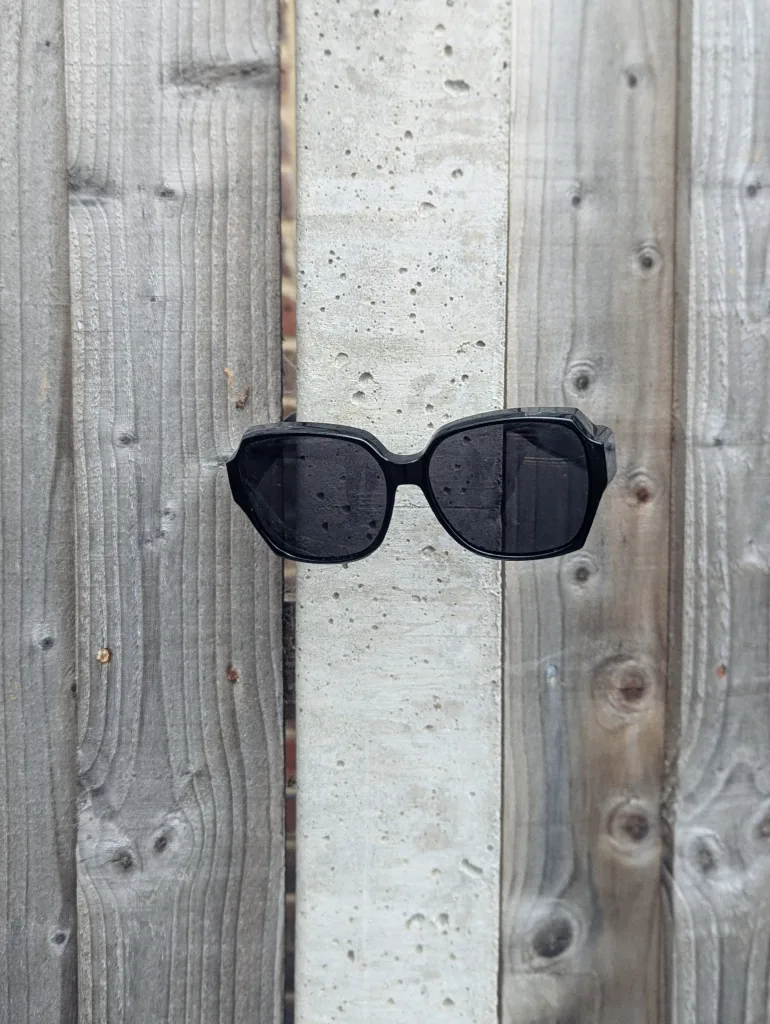 Black sunglasses balanced on a light grey concrete post between weathered wooden fence panels.