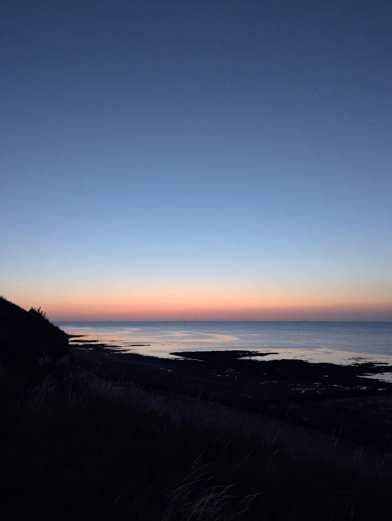 Orange twilight glow on the horizon over a calm sea, viewed from a silhouetted grassy cliff edge under a deep blue sky.