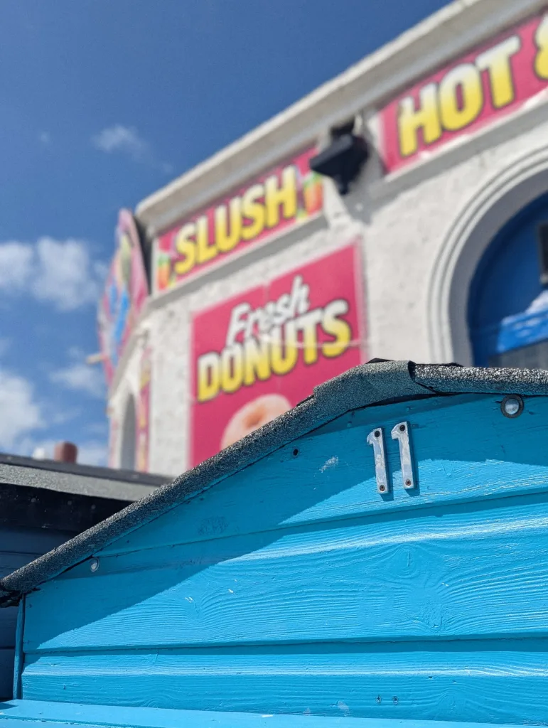 A bright blue beach hut gable with the silver number 11, set against blurred seaside snack signs for slush and donuts under a sunny sky.