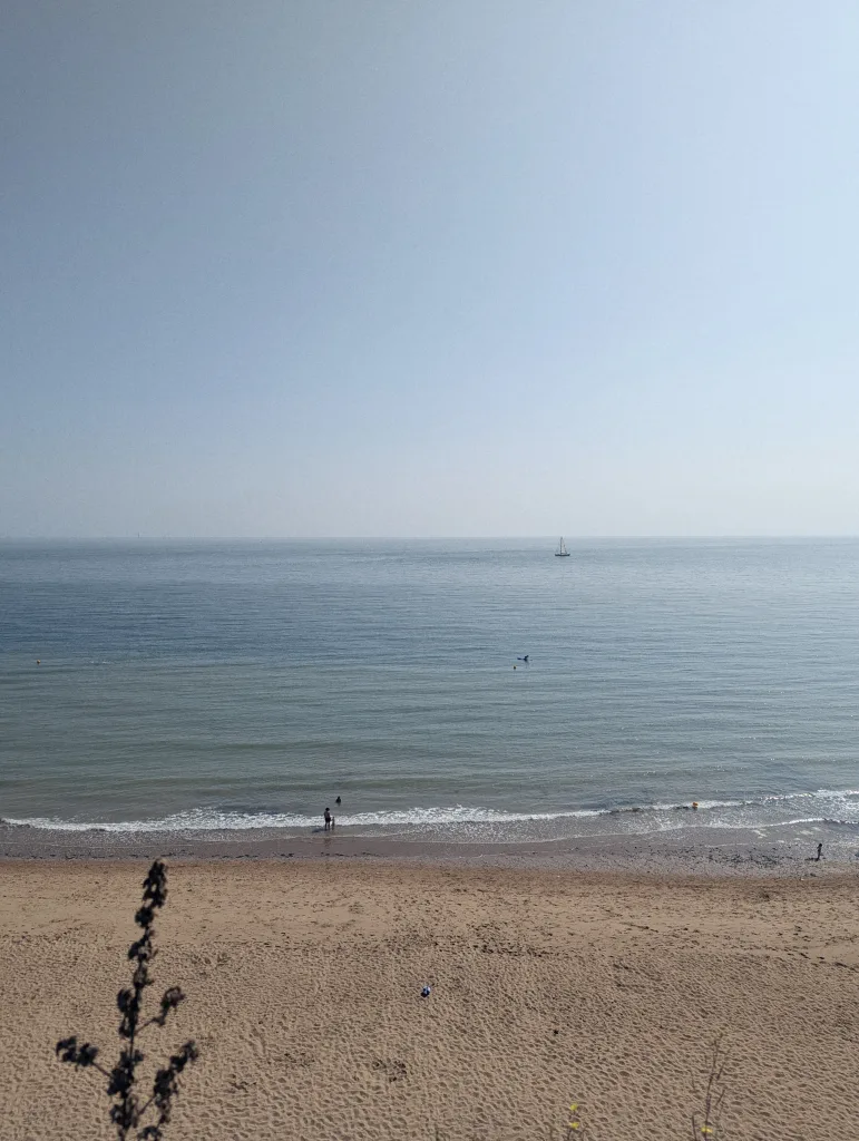 A sandy beach meets the calm blue sea with small waves under a clear pale sky, featuring a lone sailboat on the distant horizon.