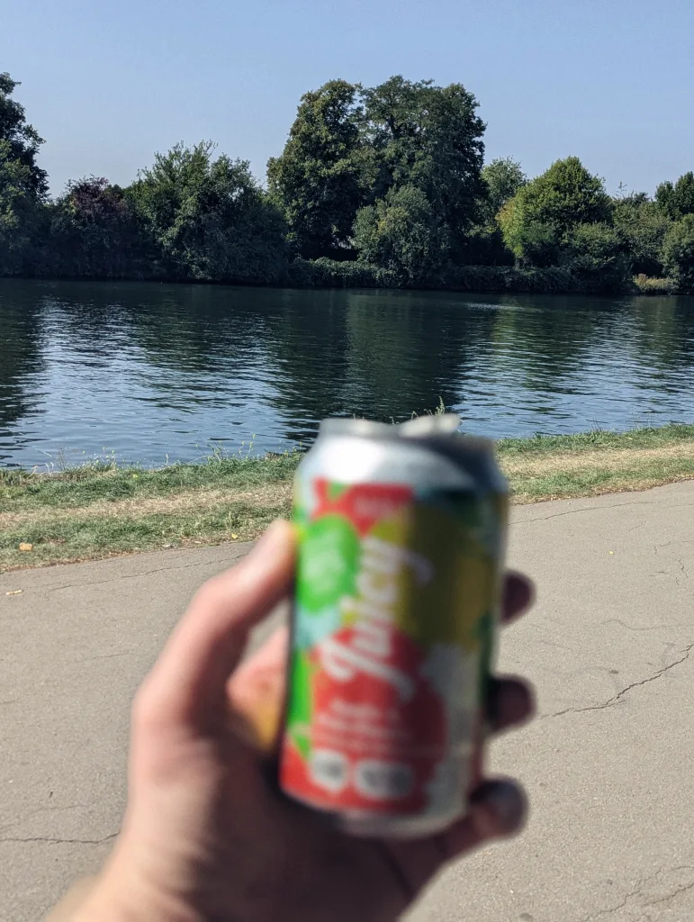 A hand holds a blurred beverage can in front of a wide river and a dense line of green trees under a clear blue sky.