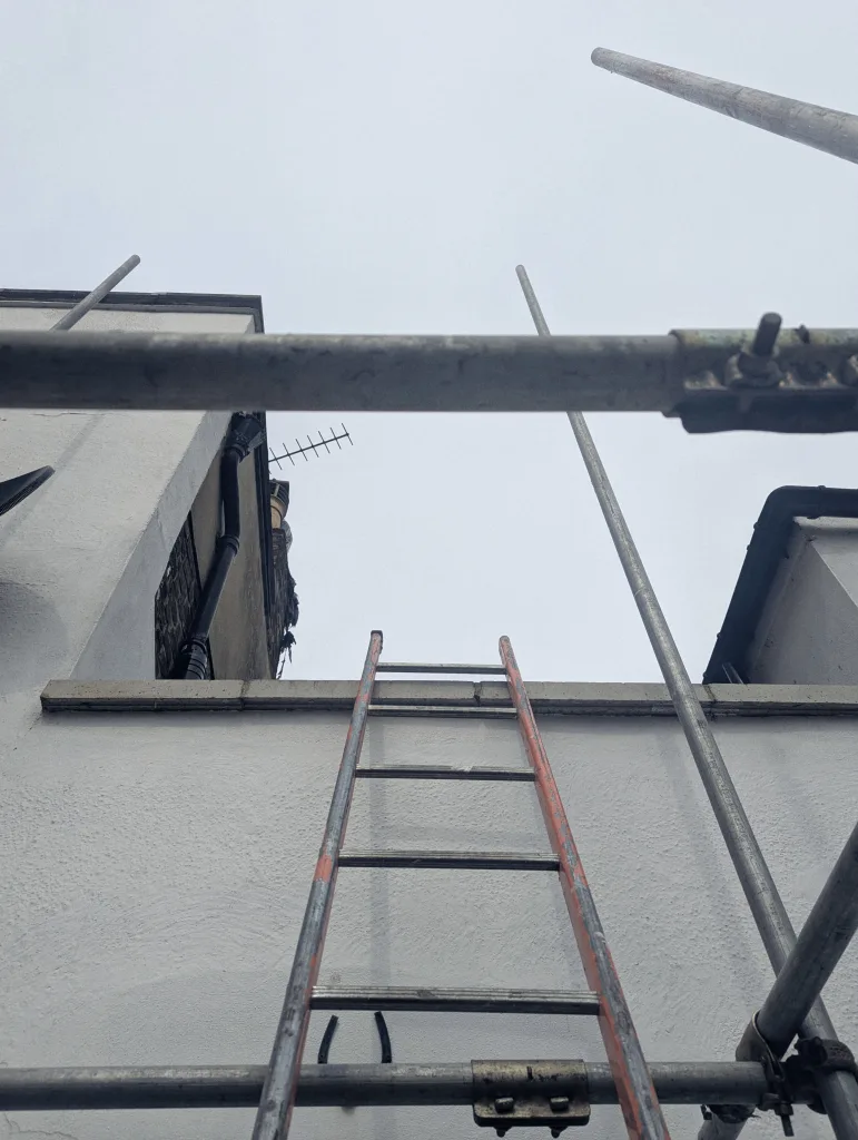 An orange ladder leaning against a white building wall, framed by grey metal scaffolding pipes looking up towards a pale sky.