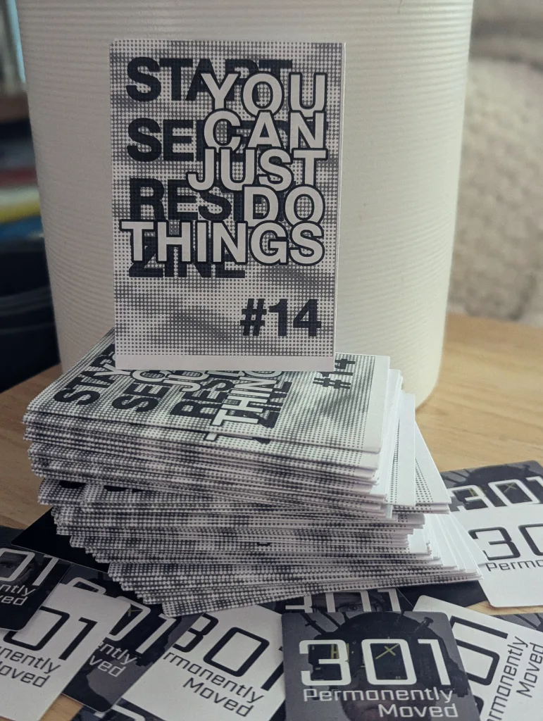 A stack of halftone zines reading YOU CAN JUST DO THINGS #14 on a wooden table beside 301 Permanently Moved stickers and a white pot.