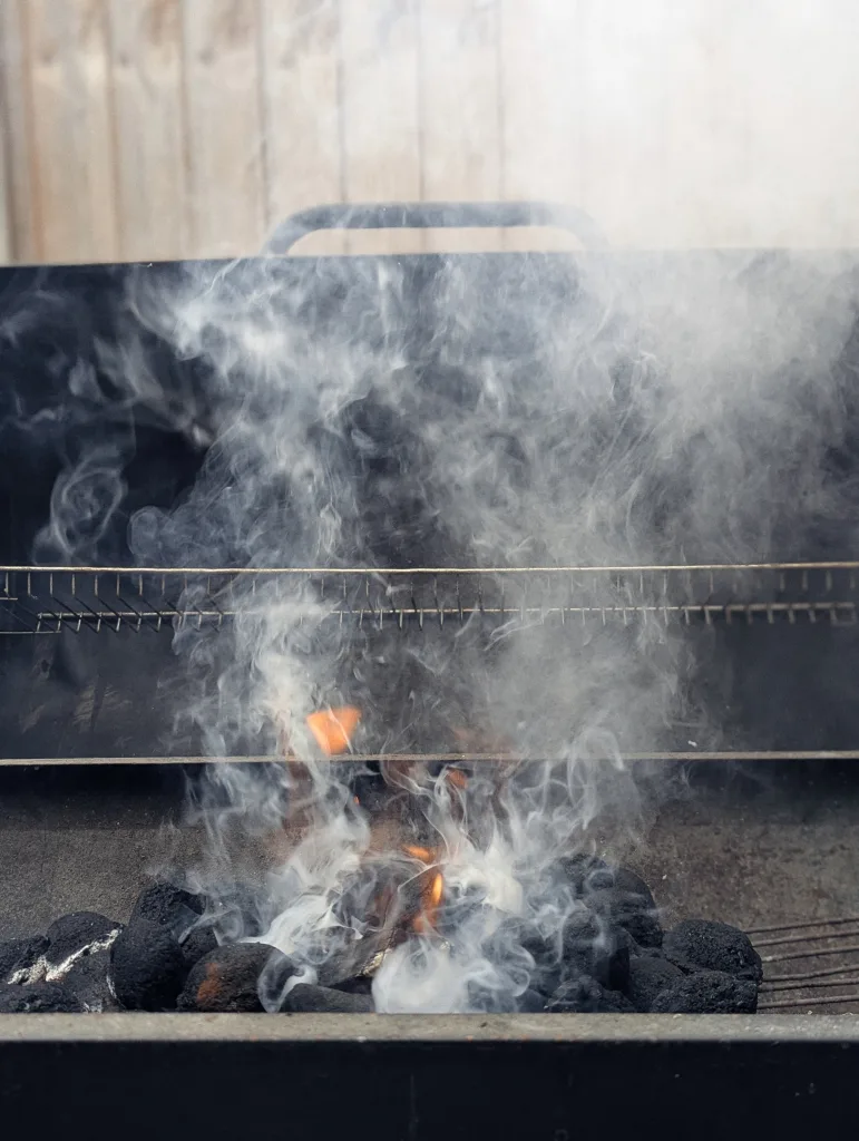 Thick white smoke and orange flames rising from glowing charcoal briquettes in a black metal barbecue grill.