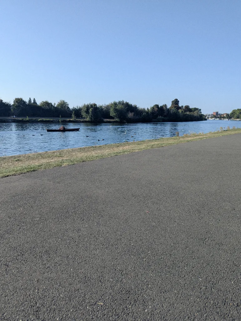 A person in a rowing boat on a wide river, seen from across a paved path and grassy bank under a clear blue sky.