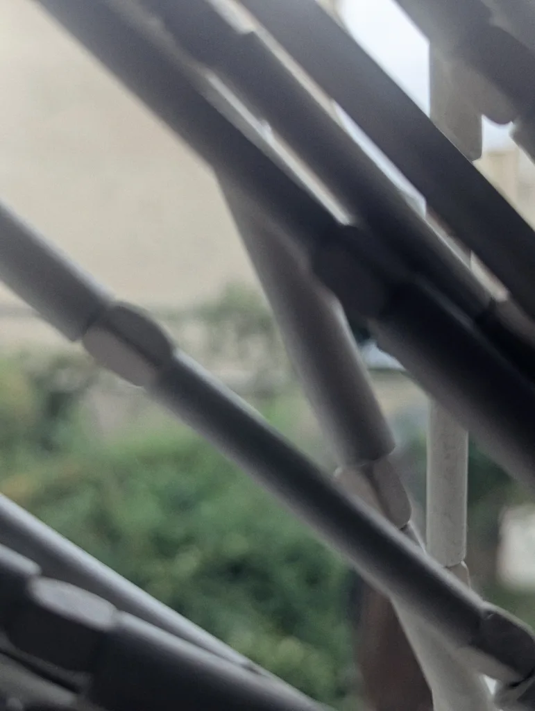 Abstract close-up of intersecting white metal rods against a blurred background of green bushes and a beige building.