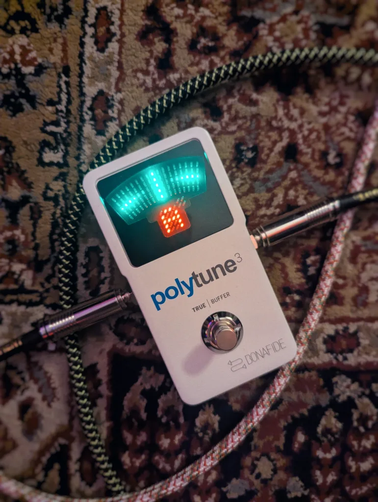White PolyTune 3 guitar tuner pedal with glowing green and red lights on a patterned rug, surrounded by coiled braided instrument cables.