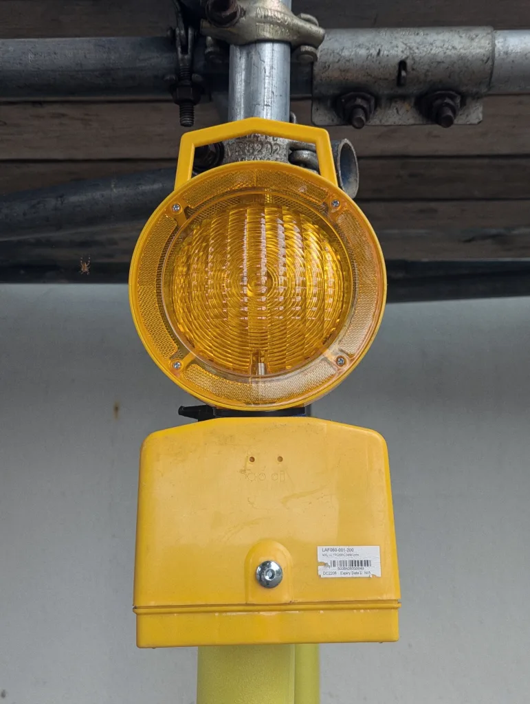 Yellow scaffolding hazard lamp with a circular amber lens attached to a metal pole. A small spider hangs on a web to the left.
