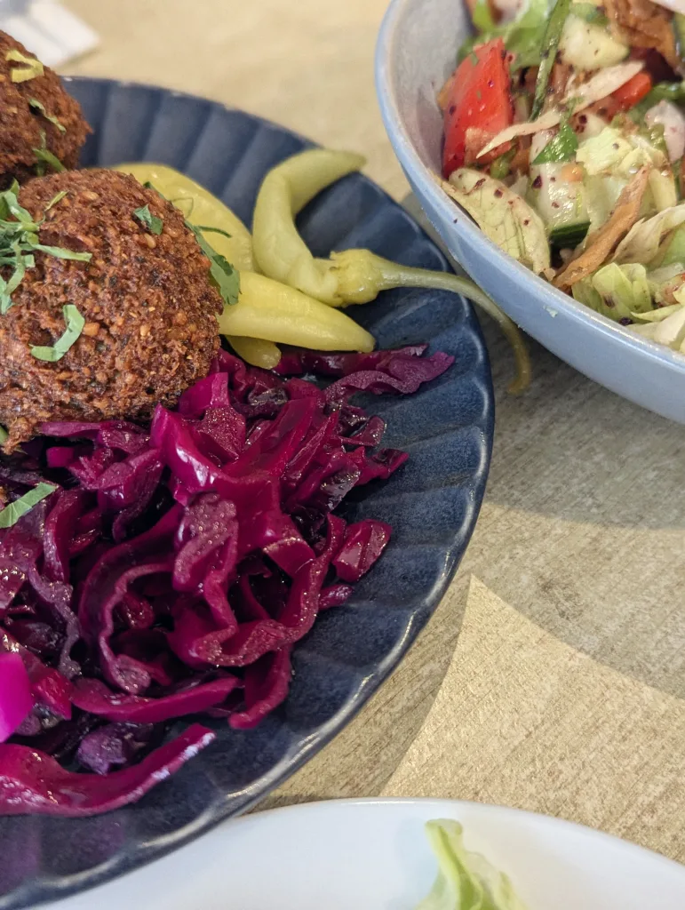 Crispy falafel with vibrant pickled red cabbage and yellow peppers on a dark blue plate beside a bowl of fresh fattoush salad.