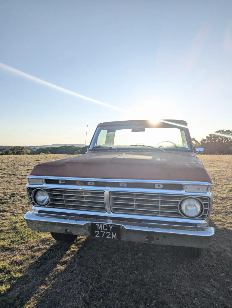 A vintage rusted red Ford truck parked in a grassy field at sunset, with bright sun flare over the cab and a clear blue sky.