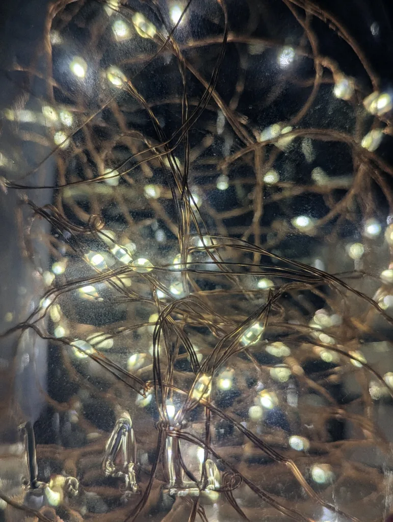 Tangled copper wire fairy lights glowing warm white inside a glass jar, creating soft light reflections.