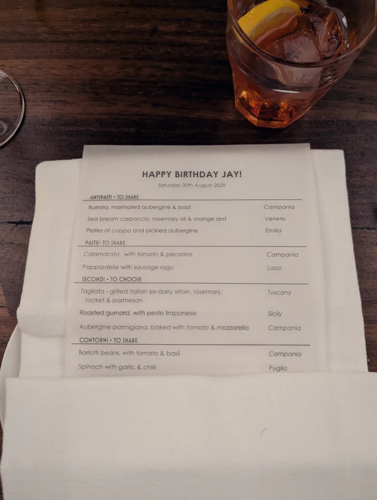 A personalized HAPPY BIRTHDAY JAY! dinner menu on a white napkin next to an orange cocktail with ice and a lemon slice on a dark wood table.