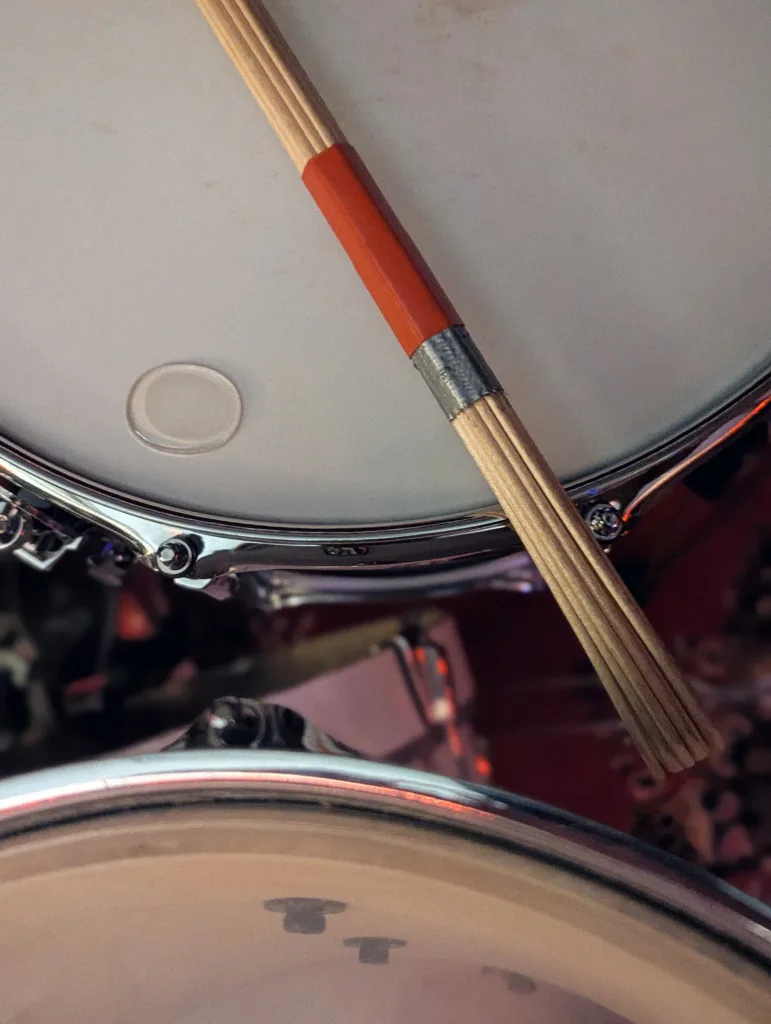 A wooden bundle drumstick with red tape resting on the white head of a snare drum.