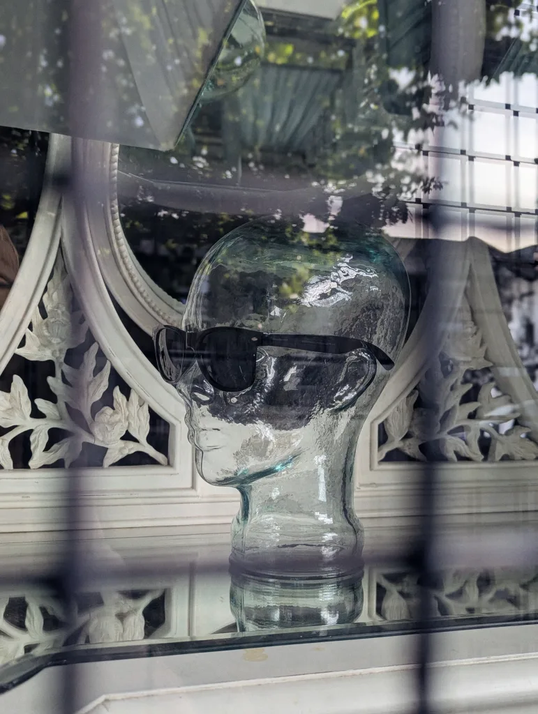 A clear glass head mannequin wearing black sunglasses in a window display, reflecting leafy trees and ornate white woodwork.